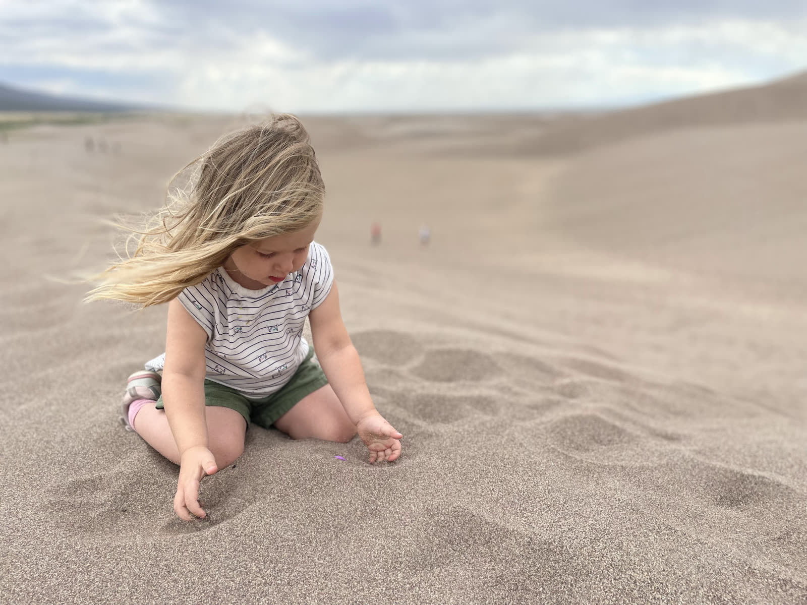 A child sitting on the Great Sand Dunes, hair lifting in the breeze, hands gently exploring the sand
